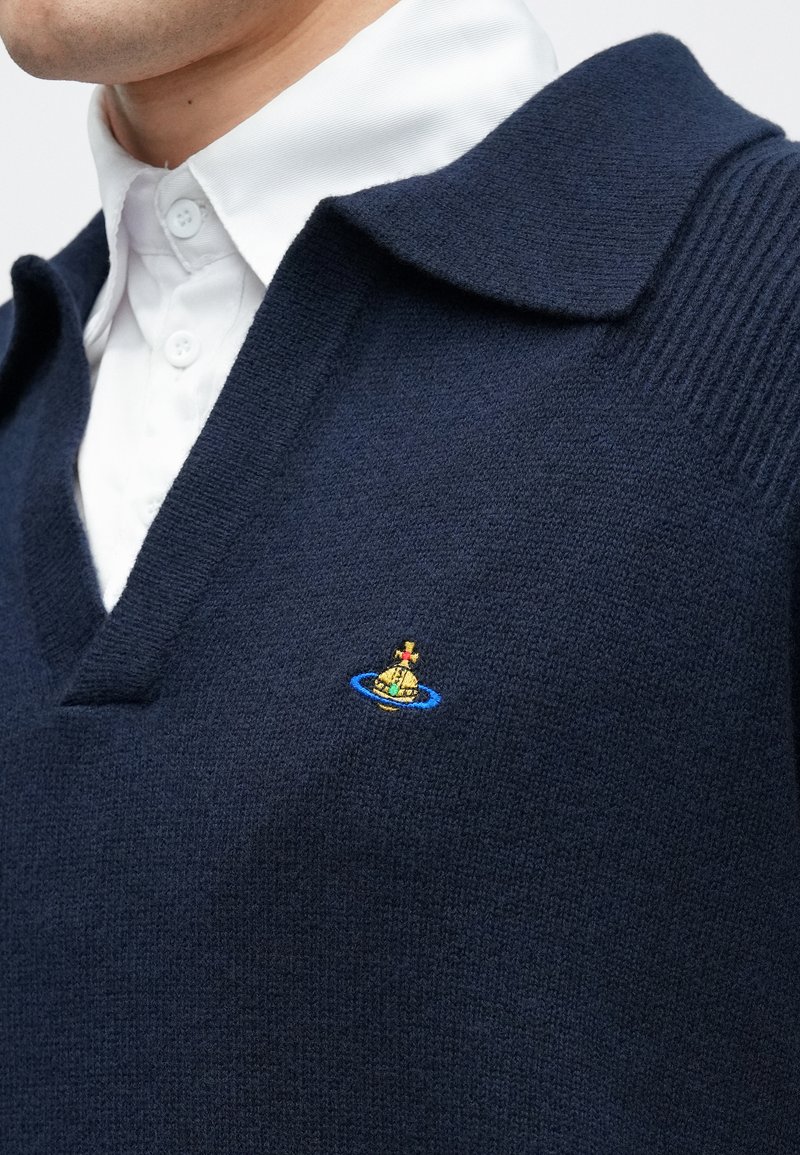 Homme portant un gilet en tricot bleu marine avec un petit logo orbe multicolore, superposé à une chemise blanche boutonnée à col.