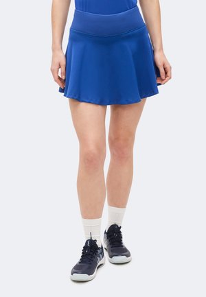 Γυναίκα που φοράει μπλε αθλητική φούστα, λευκές κάλτσες μέχρι τον αστράγαλο και μαύρα αθλητικά παπούτσια, στέκεται σε ουδέτερο φόντο.