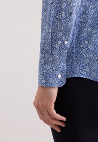 Camicia blu con motivo paisley e bottoni bianchi, dotata di polsini e una vestibilità strutturata. Tessuto liscio con intricati disegni floreali.