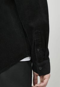 Chemise en velours côtelé noir avec des nervures verticales, des manches longues et des poignets à boutons. Présente un ourlet arrondi et un t-shirt blanc superposé.