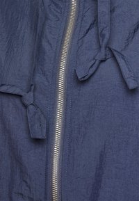 Navyblauwe lichtgewicht jas met een gestructureerde afwerking, voorzien van een zilveren rits en geknoopte strikjes aan de zijkanten voor verstelbaarheid.