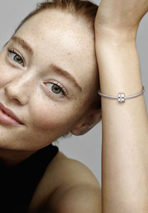 Silver snake chain bracelet with a central clasp featuring two decorative elements, worn on a wrist; light skin with freckles in the background.