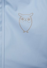 Jasnoniebieska tkanina o gładkiej fakturze z metalicznym złotym logo sowy umieszczonym na środku materiału. Projekt jest minimalistyczny i nowoczesny.