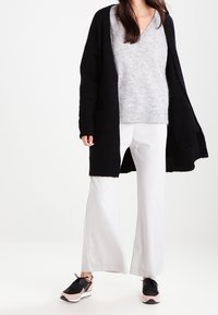 Černý dlouhý kardigan, světle šedý svetr, bílé kalhoty s širokými nohavicemi a černé tenisky s růžovými akcenty; měkké materiály a volný střih.