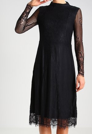 Robe noire avec de longues manches en dentelle transparente à motifs floraux, corsage texturé et ourlet en dentelle. Silhouette élégante en A.