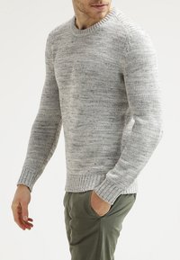 Pull gris clair en maille avec col rond, poignets et ourlet côtelés. Présente un motif texturé et une coupe ajustée. Porté avec un pantalon vert.