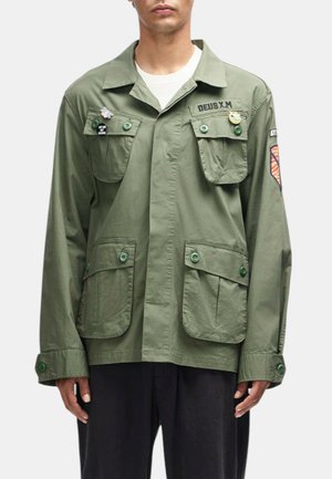 Hombre que lleva una chaqueta de estilo militar verde con grandes bolsillos con solapa, cierres de botones, parches y pins en el pecho y la manga.