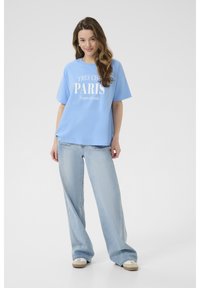Světle modré bavlněné tričko s bílým textem "TRÈS CHIC PARIS" a "Toujours à la mode." V kombinaci se světle modrými džíny, model stojící v neformální póze.