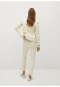 Completo su misura di colore crema con giacca aderente e pantaloni slim, realizzato in tessuto liscio, con bottoni sulla giacca e pantaloni alla caviglia.