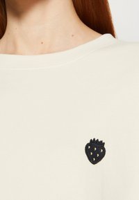 Gros plan sur une personne portant une chemise crème avec un petit écusson de fraise noire près de la clavicule.