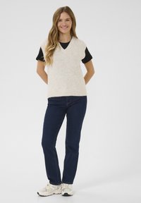Crème gebreid mouwloos top met V-hals en een gestructureerde afwerking, gedragen over een zwart T-shirt met korte mouwen. Gecombineerd met donkerblauwe jeans en lichte sneakers.