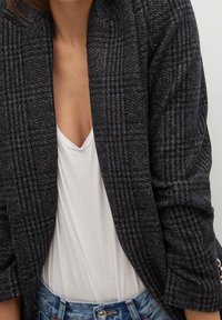 Šedý houndstooth blazer s otevřeným předním dílem, strukturovanou tkaninou a tříčtvrtečními rukávy, vrstvený přes bílé tričko a modré džíny.