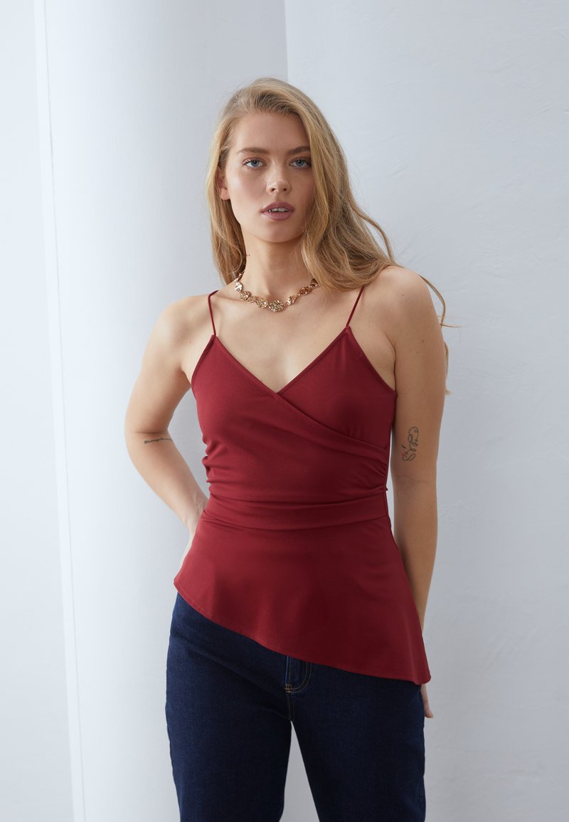 Top rosso senza maniche con design incrociato, caratterizzato da spalline sottili e un orlo svasato. Indossato con jeans blu scuro; accenti in oro con una collana.