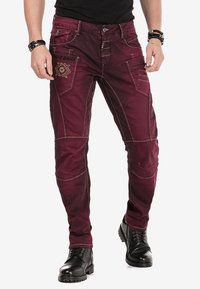 Bordeaux denim broek met contrasterende tan stiksels, voorzien van meerdere zakken en een geborduurd ontwerp aan de voorkant. Gestyled met zwarte laarzen.