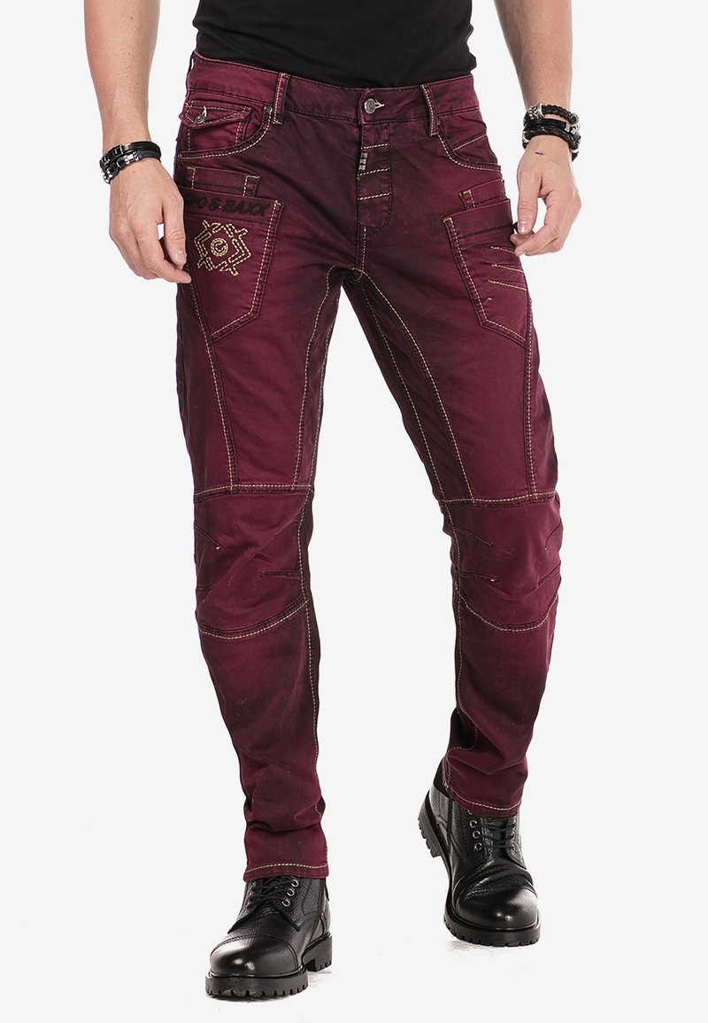Bordeaux denim broek met contrasterende tan stiksels, voorzien van meerdere zakken en een geborduurd ontwerp aan de voorkant. Gestyled met zwarte laarzen.