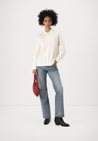 Kremfarget skjorte med krage, blå rette jeans og svarte hælboots. Modellen holder en rød håndveske.