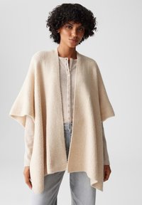 Beige, ribbed Strickponcho mit weiten Ärmeln und einem strukturierten Muster, über einem hellbeigen, knöpfbaren Oberteil getragen. Lässige Passform mit offenem Vorderteil.