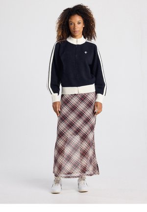 Maglione blu navy con zip, strisce bianche e polsini a coste, abbinato a una lunga gonna a quadri con toni rosso intenso e beige, indossato con sneakers bianche.