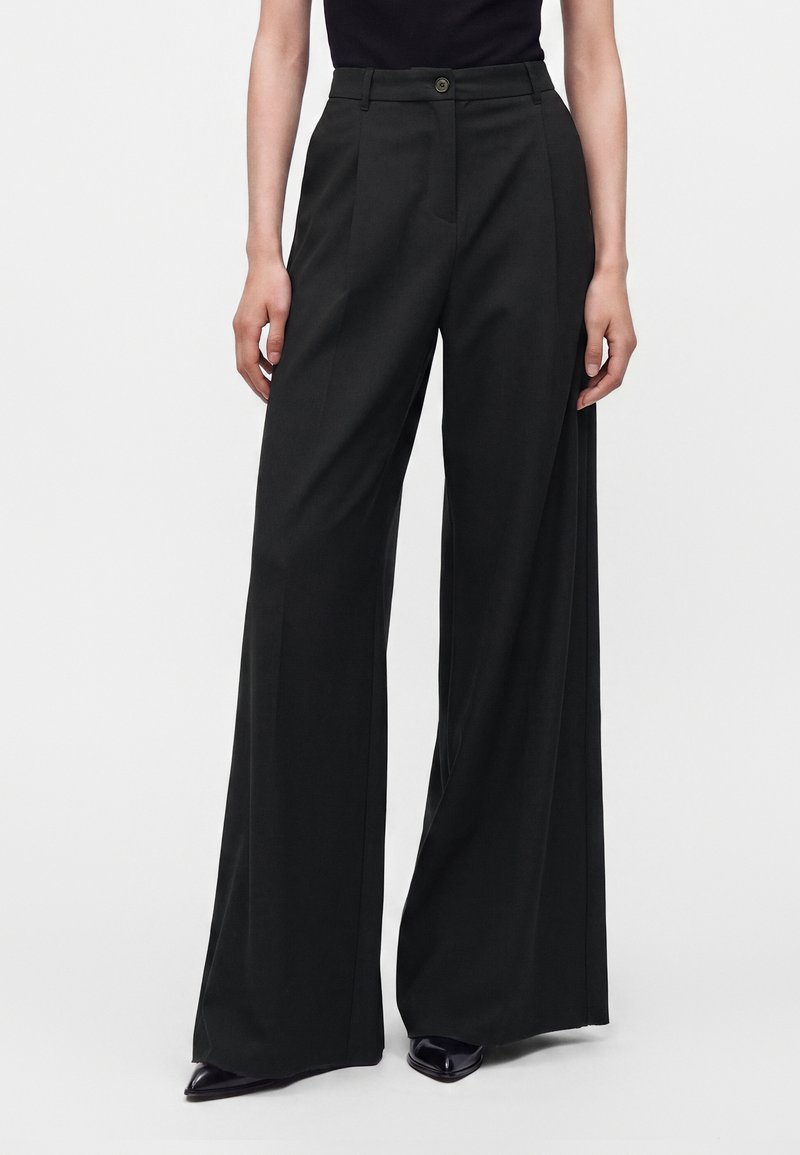 Femme portant un pantalon large noir taille haute et des chaussures noires à bout pointu, debout devant un fond clair uni.