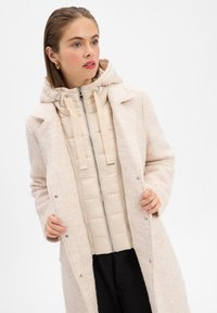 Beige gewatteerde jas met een afneembare capuchon, gelaagd onder een lichte, gestructureerde crème jas met een inkepingkraag en drukknoopsluitingen.