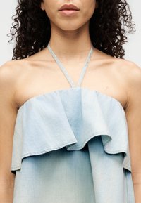 Haut en denim bleu clair avec un design à volants. Présente des fines bretelles nouées autour du cou et une texture douce. L'ourlet forme une courbe douce sous la poitrine.