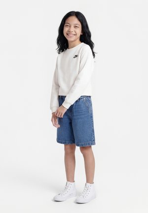 Χαμογελαστή κοπέλα με μακριά μαύρα μαλλιά φοράει λευκό φούτερ Nike, μπλε τζιν σορτς μέχρι το γόνατο και λευκά ψηλά αθλητικά παπούτσια, στέκεται σε λευκό φόντο.
