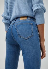 Suéter de punto azul claro con mangas enrolladas, combinado con unos vaqueros de lavado medio que tienen una etiqueta de cuero y dos bolsillos traseros.