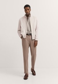 Helle rosa Bomberjacke, graues Strickhemd, beige Hose, braune Lederschuhe. Maßgeschneiderte Passform und minimalistischem Design mit Reißverschlussdetails.