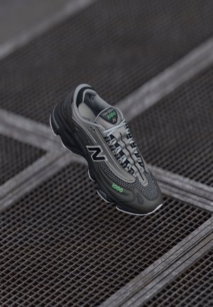 Sort og grå New Balance 1000 sneakers med grønne accenter, der svæver over et metalristsgulv.