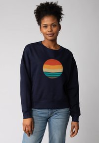 Marineblauwe sweatshirt met een ronde graphic die horizontale strepen in oranje, geel en teal toont. Relaxed fit en ribgebreide mouwen.
