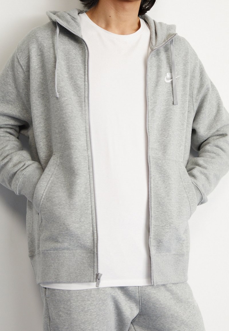 Šedá mikina na zip vyrobená z měkké látky, obsahující přední zip, nastavitelné šňůrky a boční kapsy; logo na prsou.