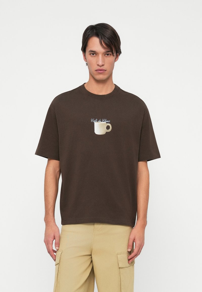 Καφέ βαμβακερό μπλουζάκι με κοντά μανίκια, που διαθέτει μια λευκή γραφική απεικόνιση φλιτζανιού καφέ και κείμενο που γράφει "Café de Choux" σε καλλιγραφική γραφή.