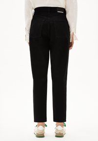 Person som bär högmidjade svarta jeans med bakfickor, en vit långärmad skjorta och vita sneakers med gröna accenter, stående med ryggen mot kameran.