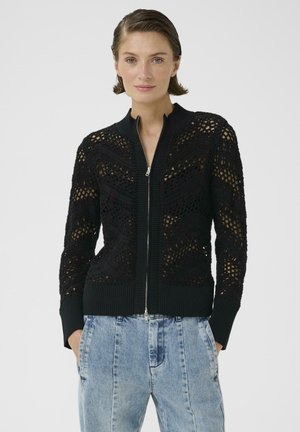 Femme portant un pull zippé noir en crochet et un jean taille haute bleu clair, les mains dans les poches, devant un fond blanc uni.