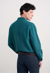 Cămașa din in teal cu mânecă lungă are un guler cu nasturi și mansete ajustabile, fiind combinată cu pantaloni închiși la culoare și o curea împletită maro.