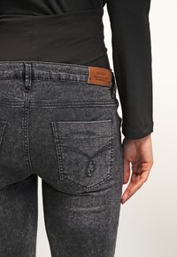 Jeans in denim nero con una finitura testurizzata, dotati di un patch in pelle marrone sulla cintura e tasche posteriori angolate con dettagli cuciti.