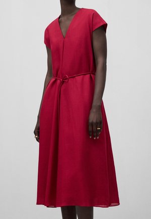 Kvinde iført en knælængde, ærmeløs rød kjole med V-udskæring og tyndt bælte i taljen, stående foran en ensfarvet grå baggrund.