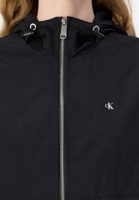 Czarna kurtka z kapturem, wykonana z lekkiego materiału, z metalowym suwakiem, regulowanymi sznurkami oraz małym białym logo na klatce piersiowej.