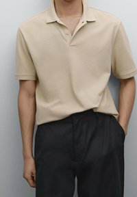 Beige pikétröja med krage, korta ärmar och mjukt material. Matchat med mörka byxor, vilket ger en avslappnad och modern look.