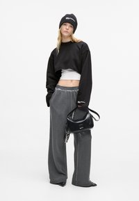 Črn skrajšan pulover čez bel top, sivi široki hlače in sijoča črna torbica z logotipom na traku. Manekenka nosi črno kapa.