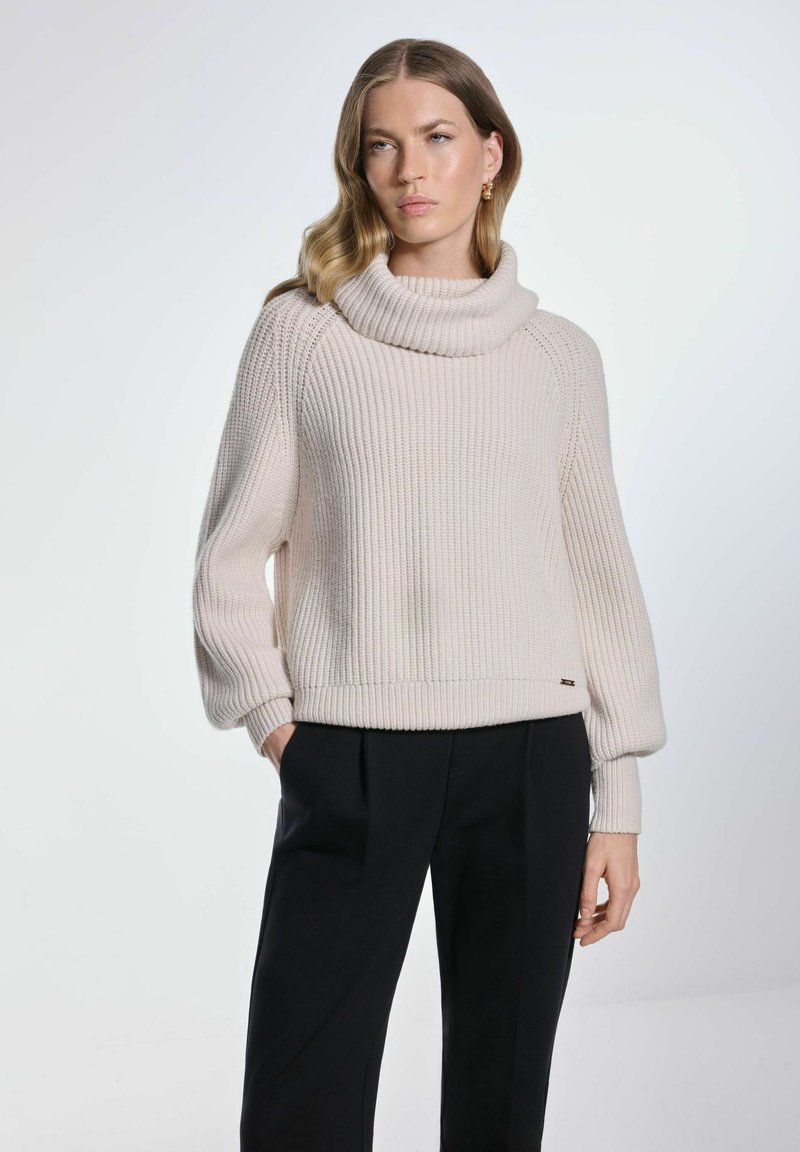 Jasnobeżowy sweter ribbed z dużym kołnierzem typu cowl oraz długimi, lekko bufkowymi rękawami, w połączeniu z czarnymi spodniami. Wyraźne detale przeszycia.