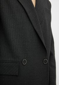 Blazer nero a righe verticali doppio petto con una finitura testurizzata. Presenta bottoni in tono argentato e rever a picco. Include una tasca laterale.