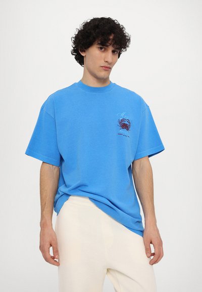 Blue short-sleeve t-shirt in cotton, featuring a round neckline and front graphic design. Worn with light-colored trousers.