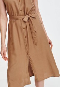 Beżowa sukienka zapinana na guziki z przewiązanym w pasie paskiem, wykonana z lekkiego materiału. Ma długość do kolan i gładką fakturę z delikatnym połyskiem.