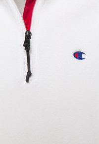 Weißer Fleece-Pullover mit einem Viertel-Reißverschluss, ausgestattet mit einem roten Reißverschluss und schwarzen Beschlägen. Gesticktes Logo in Blau und Rot auf der linken Brust.
