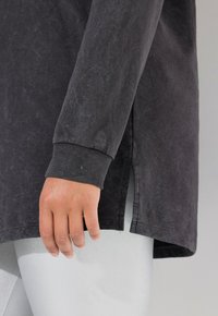 Dunkelgraues Langarmshirt mit strukturiertem Finish, gerippten Bündchen, Seitenschlitzen und sichtbaren Nähdetails. Hand ruht auf dem Stoff.
