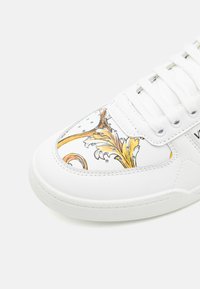 Weißer Sneaker mit einem glatten Lederobermaterial, das detaillierte goldene und schwarze Blumenmuster auf der Zehenkappe und den Seitenwänden aufweist.