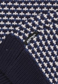 Marineblauwe en witte gebreide sjaal met een houndstoothpatroon. Ribbelstructuur aan de uiteinden, met een zwart label met witte tekst.