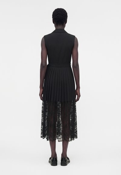 Άτομο με μαύρο αμάνικο φόρεμα με πλισέ φούστα και διαφανή δαντέλα στο πάνω μέρος, στέκεται πλάτη σε μονόχρωμο λευκό φόντο.