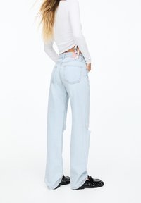 Jeans de pierna ancha en azul claro con detalles desgastados y una etiqueta rosa; llevados con una blusa ajustada de mangas largas blanca y mocasines negros con tachuelas.
